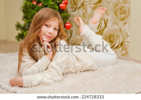 Girl lying on the carpet, looking at the camera and smiling. A child in a warm white sweater. Christmas tree. Teen in New Year clothes.