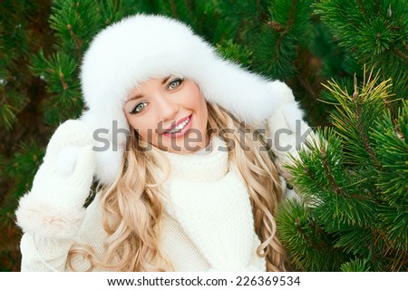 Christmas poster. Young smiling woman with fur white hat, woolen mittens, sweaters in the winter fir forest. Fir branches.