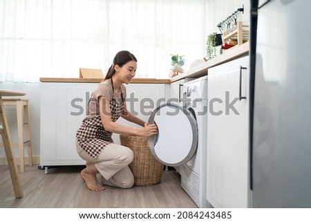 Asian beautiful woman put dirty clothes to washing machine in house. Attractive girl wear apron sit on floor, feel happy to loads laundry in washer appliance at home. Domestic-House keeping concept. Foto stock © 