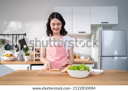 Asian young attractive woman making sandwich for breakfast in morning. Beautiful girl wear apron enjoy eat clean vegetables healthy food for health care in house. Diet lose weight and foods concept. Foto stock © 