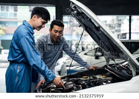 Asian automotive mechanic explain car condition to client in garage. Vehicle service manager male work in mechanics workshop point at vehicle part to customer man for maintenance and repair car engine Foto stock © 