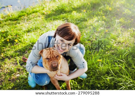 laughing happy young woman in denim overalls hugging her red cute dog Shar Pei in the green grass in sunny day, true friends forever, people pets concept