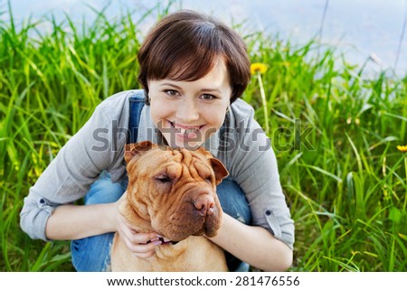 portrait of laughing happy young woman in denim overalls hugging her red cute dog Shar Pei in the green grass in sunny day, true friends forever, people pets concept