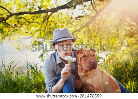 happy young woman in denim overalls and hat with red cute dog Shar Pei sitting in the lawn in sunset light and blowing on a dandelion flowers, best friends forever, people concept