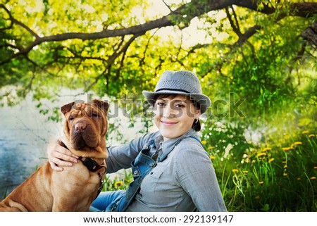 Happy woman in denim overalls and hat with her dog Shar Pei sitting in the meadow near the lake at sunset, true friends forever, people pets concept