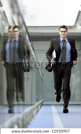 Businessman with laptop under his arm runs along modern office building corridor