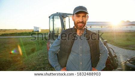 Portrait of young Caucasian handsome happy man farmer standing in field and smiling to camera. Big tractor on background. Cheerful male worker in agricultural farm. Sunlight. Agriculture farming.