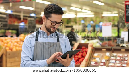 Close up portrait of happy Caucasian male worker in glasses standing in supermarket and typing on tablet. Young joyful man food store assistant at work tapping on device indoors. Retail concept Imagine de stoc © 