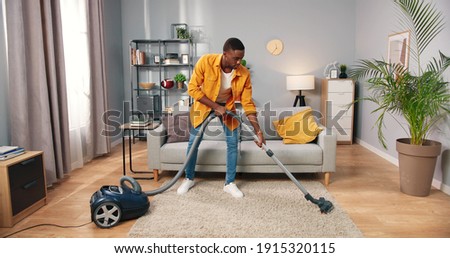 African American busy handsome young guy vacuuming cozy living room doing housework, male cleaning house using vacuum keeping home clean, housekeeper, everyday life concept
