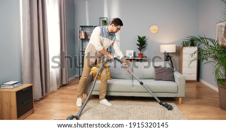 Caucasian handsome serious young man vacuuming carpet cleaning modern apartment, clean up, housekeeper, guy doing domestic tasks and cleans living room using vacuum cleaner, householding