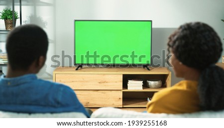 Back view of young African American family couple watching TV with chroma key while resting at home sitting on sofa in room. Wife and husband spending time together watch television with green screen