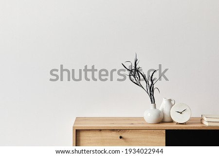 Minimalist concept of living room interior at elegant apartment with wooden commode, leaf in ceramic vase, clock and elegant personal accessories in modern home decor. Copy space. Template. .