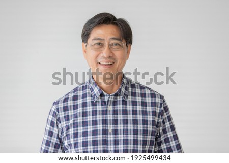 Portrait close up shot of middle aged asian male model with short black hair wearing blue plaid shirt with stand smiling in front of white background.