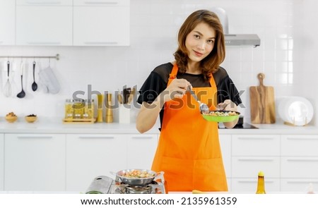 Mom in orange apron in home kitchen happily smile and pride with food on spoon she picking up from flying pan as success to learn family cooking for kid. 商業照片 © 