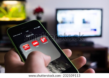 BUCHAREST, ROMANIA - NOVEMBER 21, 2014: Photo of hand holding an iphone with news apps on screen and tv set in the background with news channel