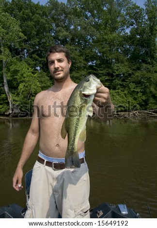 Young man holding a large mouth bass that he has just caught out of a river.