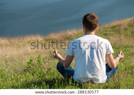 Young man sits on the top of the hill viewing wide Dnister river. Yoga meditation