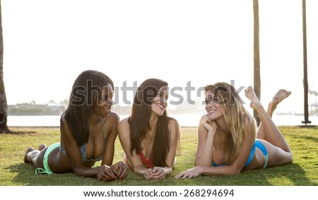 Three women lying on their belly, on the grass in a park. Backlit, looking at each other.