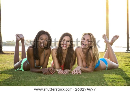 Three women lying on their belly, on the grass in a park. Backlit.