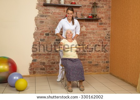 Senior Woman Sitting On A Chair And Doing Exercise With A Stick During Physiotherapy, Looking At Camera