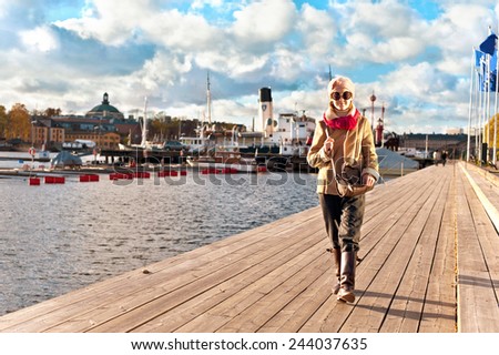 Cute smiling young stylish and trendy girl walking on pier in Stockholm on cloudy sky background. Autumn outdoors.