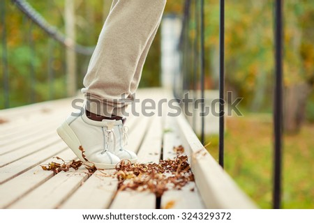 Woman feet in white shoes standing tiptoe on hanging bridge in autumn park. Side view. Outdoors fall season multicolored horizontal image.