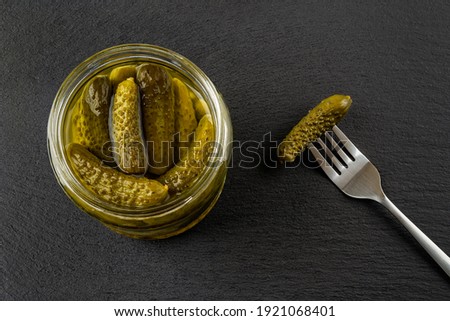 Open glass jar of tasty pickled gherkins and one more on the spoon against black slate background. Whole green cornichons marinated with dill, garlic and mustard seeds. Crunchy baby pickles. Top view. Photo stock © 