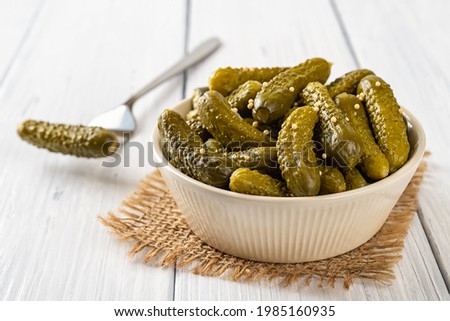 Crunchy pickled gherkins in a beige ceramic bowl and one on the fork on a white wood table. Whole green small cucumbers marinated with dill and mustard seeds. Delicious baby pickles. Canned vegetables