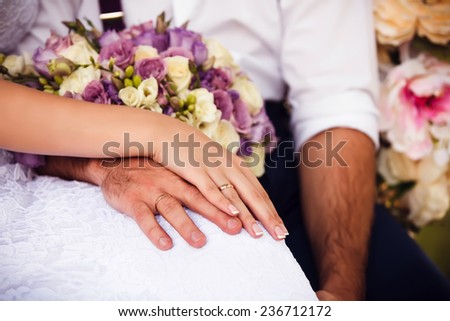 Bride and groom\'s holding each other\'s hand with wedding rings. Wedding bouquet of yellow, pink and purple roses lying on their knees. Photos made in warm colors