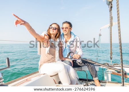 Beautiful couple of lovers sailing on a boat - Young attractive man holding rudder of a yacht and looking far away - Two fashion models posing on a sailing boat at sunset