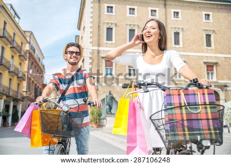Happy couple riding bikes in the city - Young man and woman with shopping bags and she speaks at mobile phone