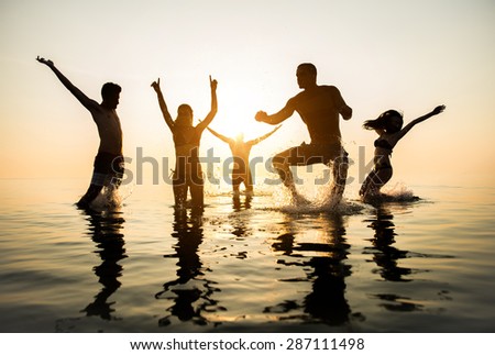 Group of happy friends jumping in to water at sunset - Silhouettes of active people dancing and having fun on the beach on vacation