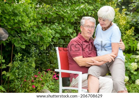 An romantic elderly couple sitting outside, the wife is sitting on the husband and looking down