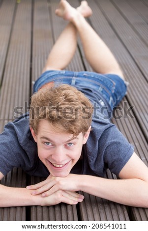 A young blonde male model is resting on his hands, lying on a brown wooden surface and laughing