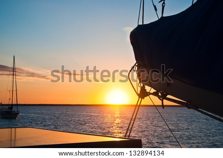 Sun setting over the ocean with the boom and mainsail of a sailboat in the foreground and another sailboat anchored in the distance copy space available