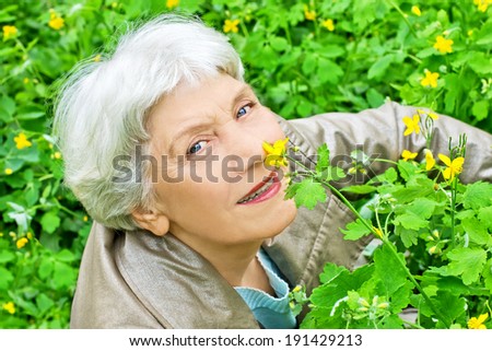 Happy elderly woman  senior sitting on a glade of yellow flowers in spring and smelling a flower