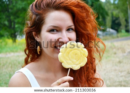 Beautiful young redhead woman smiling happily with a big flower shaped yellow lollipop