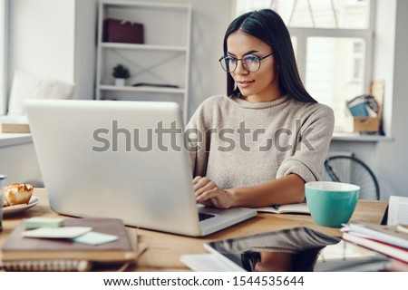 Photo of Beautiful young woman in casual clothing using laptop and smiling while working indoors