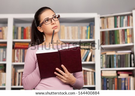 Thoughtful student. Beautiful young woman holding a book and looking away while standing at the library