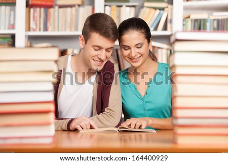 Studying together. Cheerful young man and woman sitting close to each other at the library desk and reading book