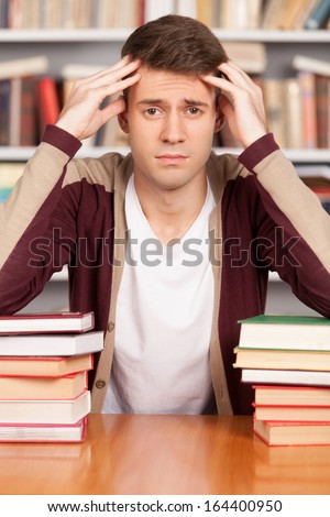 Tired of studying. Tired young man holding his head in hands and leaning at the book stack while sitting at the library desk
