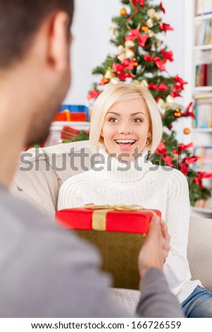 Is it for me? Cheerful young woman sitting on the couch and receiving a gift box from her boyfriend sitting close to her
