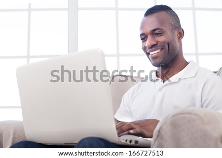 Working at home. Cheerful African man using computer and smiling while sitting on the chair