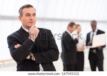 In search of the new ideas.  Thoughtful mature man in formalwear holding hand on chin and looking away while his colleagues standing on background