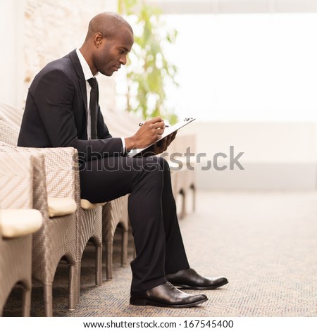 Making notes. Side view of confident young African businessman writing something in his note pad while sitting on the chair