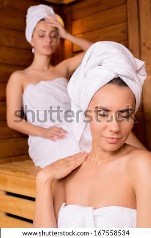 Relaxing in sauna. Two attractive women wrapped in towel relaxing in sauna and keeping eyes closed