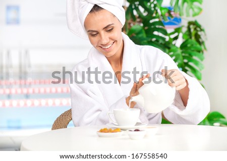 Pouring herbal tea. Attractive young woman in bathrobe pouring tea to the cup and smiling while sitting in front of swimming pool
