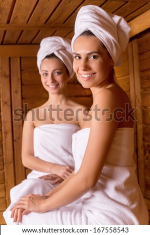 Spending time in sauna. Two attractive women wrapped in towel sitting at sauna and smiling to you