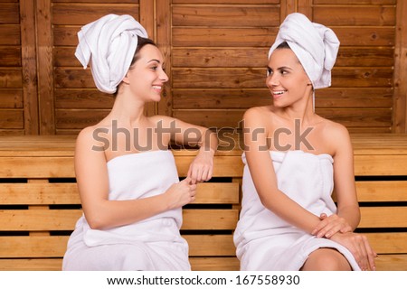 Spending time in sauna. Two attractive women wrapped in towel talking to each other while relaxing in sauna