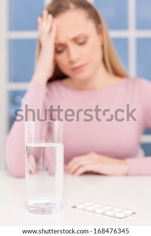Feeling headache. Depressed young woman holding head in hand while glass of water and medicines laying on foreground